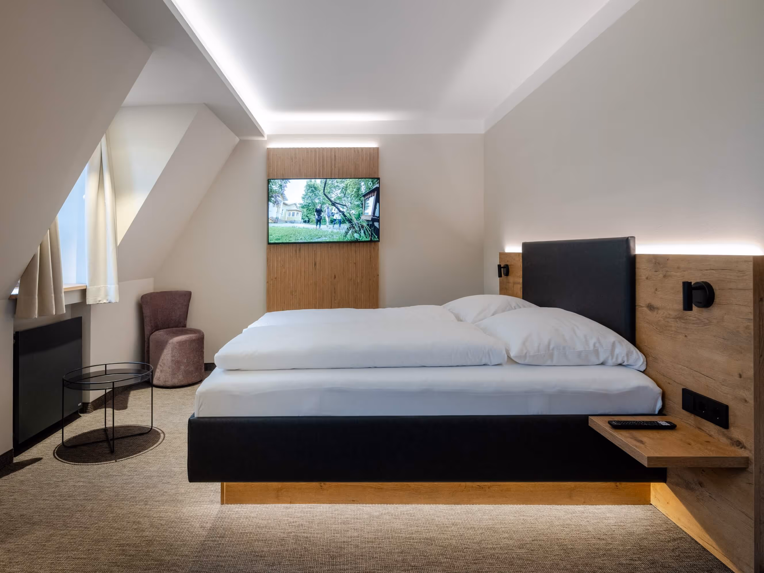 Minimalist junior suite at the Nashi Rooms Hotel near the Krämerbrücke in Erfurt with double bed, black and wooden accents, wall-mounted television, small round table and purple chair near the window.