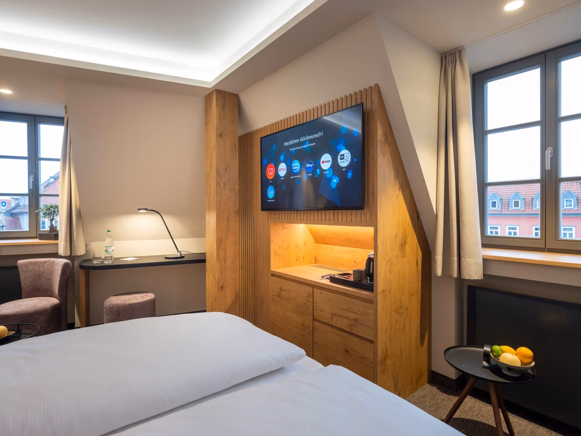Nashi Rooms hotel room with double bed, wooden TV cabinet with flat-screen TV, two windows with a view of the Krämerbrücke in Erfurt and a sitting area with a desk and chair.