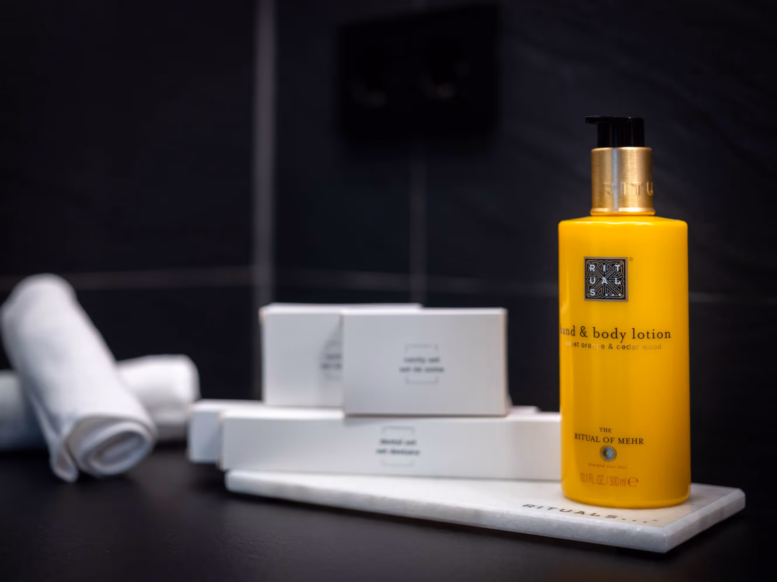 Eine gelbe Flasche Ritual of Mehr Hand- und Körperlotion von Rituals steht neben zusammengerollten weißen Handtüchern und weiteren Produkten auf einer schwarzen Oberfläche im Badezimmer des Nashi Rooms Hotelzimmers in Erfurt.