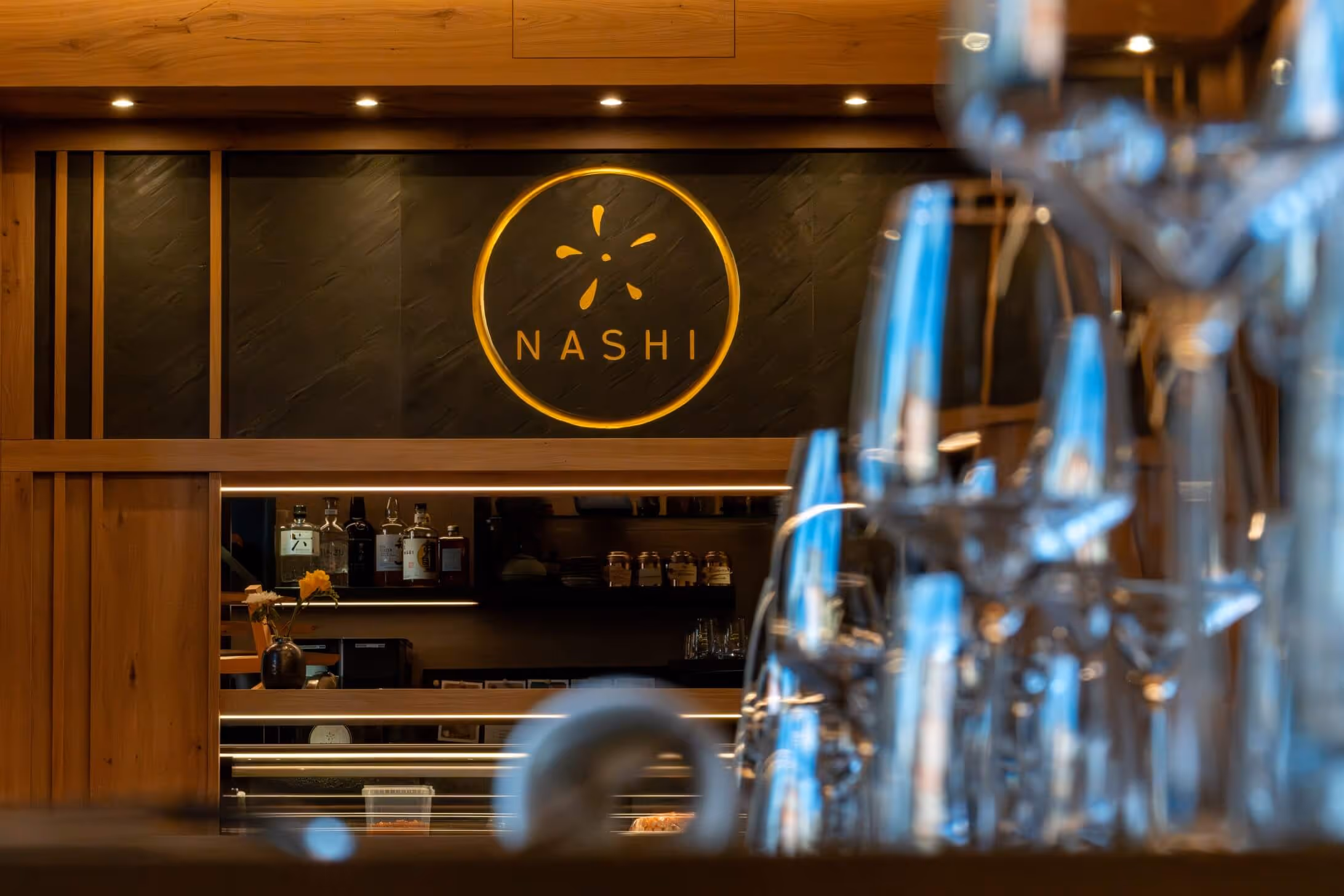 Interior of Nashi Dining in Erfurt with restaurant bar and a round NASHI logo on a black wall, glasses can be seen in the foreground.
