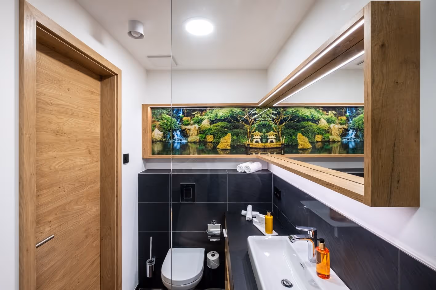 Modernes Badezimmer mit Holztür, weißem Waschbecken, schwarzen Fliesen und einem großen Spiegel mit einem malerischen Naturmotiv über der Toilette.