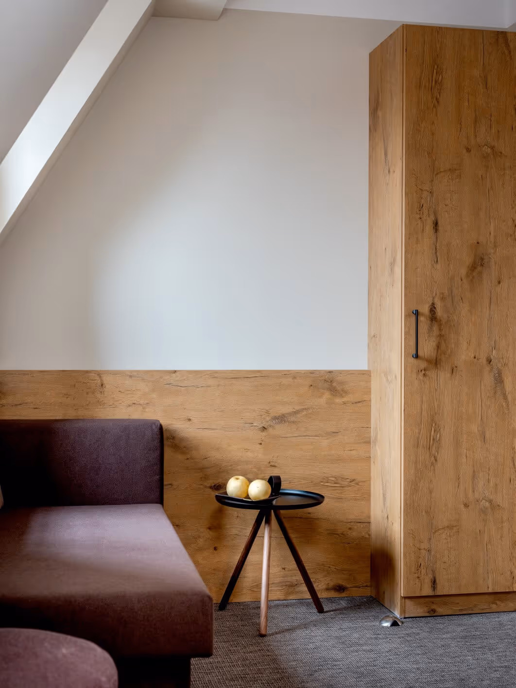 Moderne Sitzecke im Nashi Rooms Hotel in Erfurt mit lila Schlafsofa, rundem schwarzem Beistelltisch mit zwei Birnen und Holzschrank vor beiger, holzgetäfelter Wand.