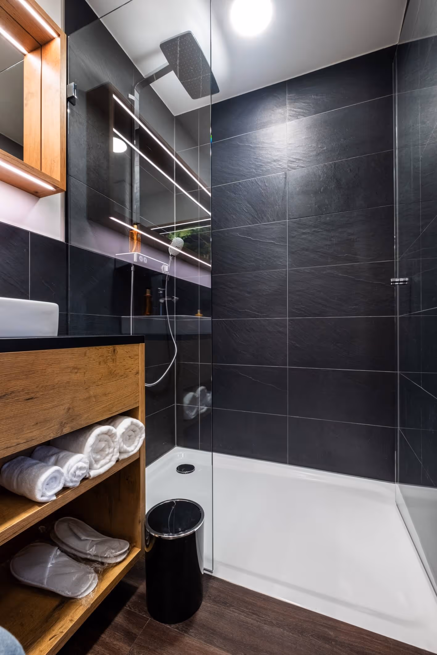 Modernes Badezimmer im Nashi Rooms Hotel in Erfurt mit dunkel gefliesten Wänden, einer verglasten Regendusche und Holzregalen in der Nähe der Krämerbrücke.