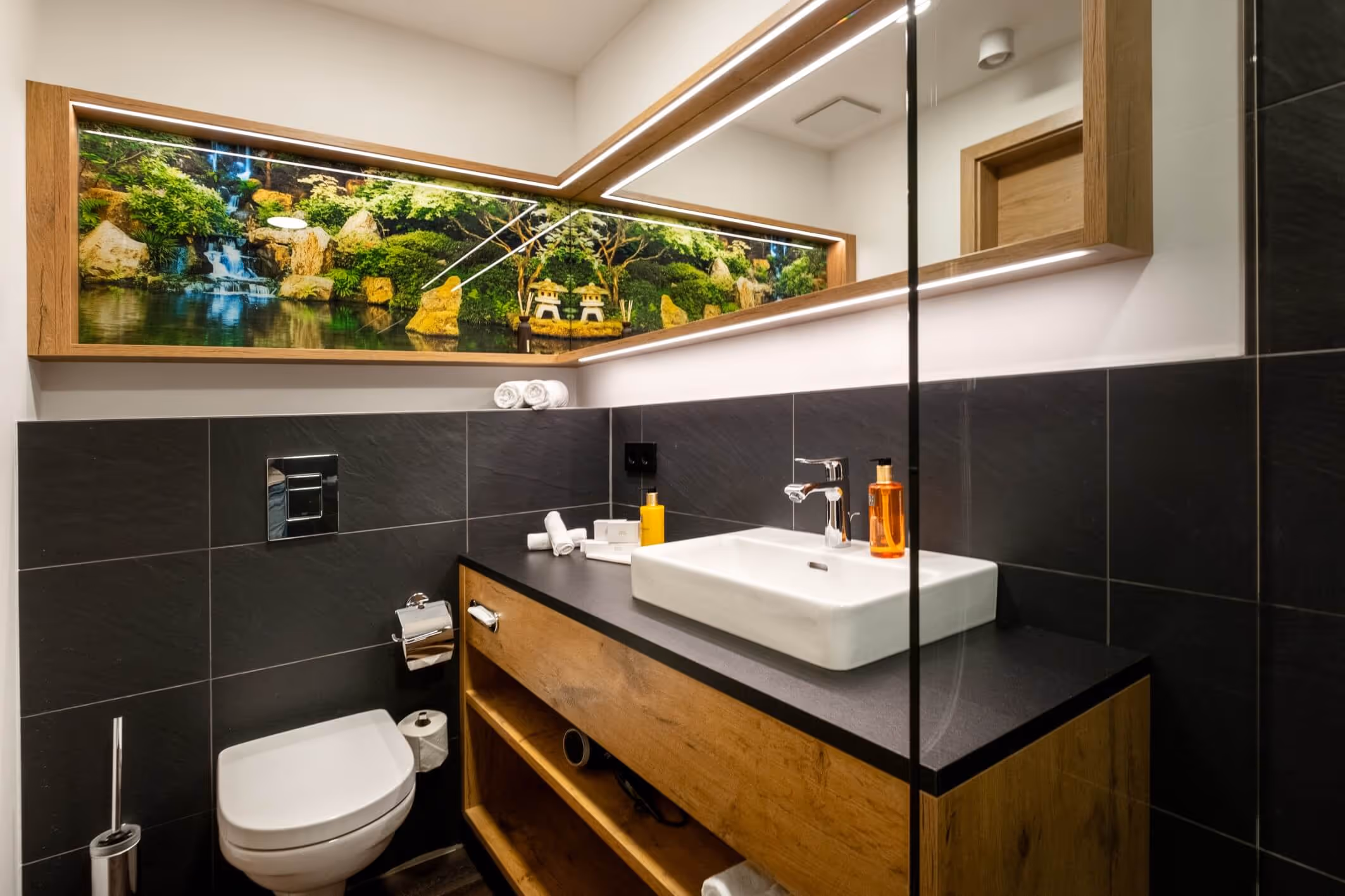 Modernes Badezimmer im Nashi Rooms Hotel in Erfurt mit weißer Toilette, schwarz gefliesten Wänden, Waschtisch aus Holz mit schwarzer Arbeitsplatte, weißen Waschbecken und einem großen gerahmten Spiegel mit einem Naturmotiv mit Wasserfall.
