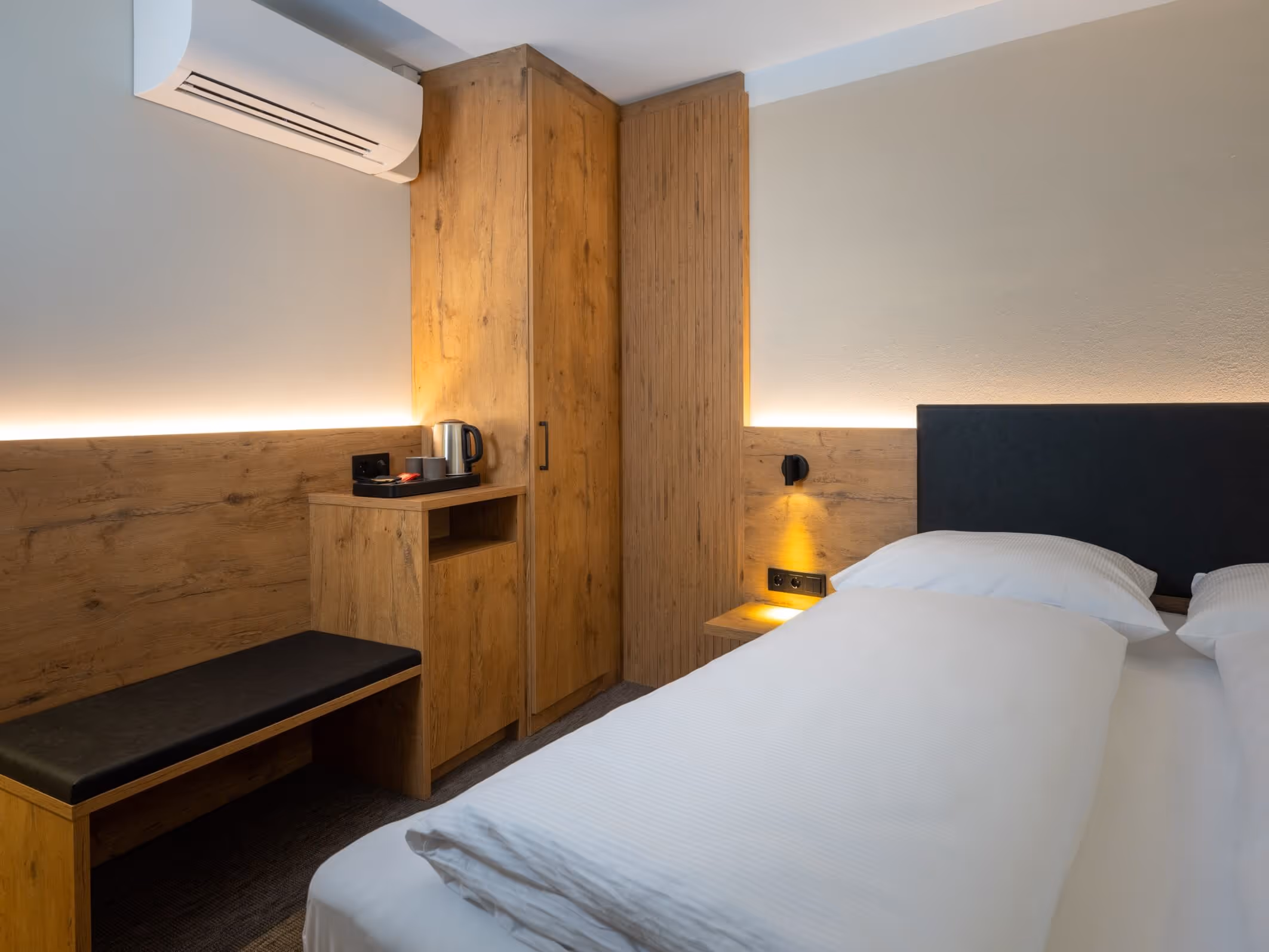 Moderne Hotelzimmerecke mit Doppelbett, Holzschrank, Sitzbank und Wasserkocher auf einem Holzschrank im Nashi Rooms Hotel im Zentrum von Erfurt.