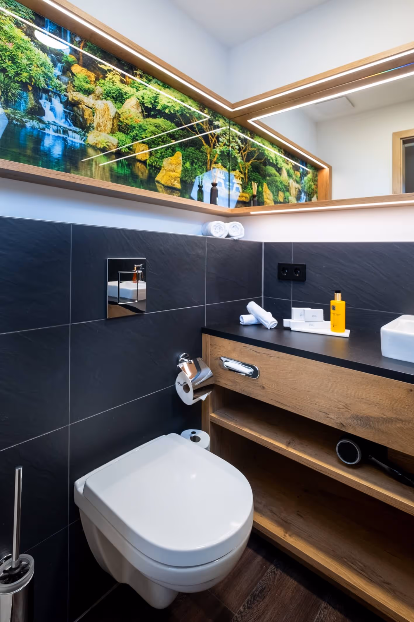 Modernes Badezimmer im Doppelzimmer, Nashi Rooms Hotel in Erfurt, mit weißer Toilette, Waschtisch aus Holz und einem großen Spiegel mit Naturmotiv, Wasserfall und Felsen.