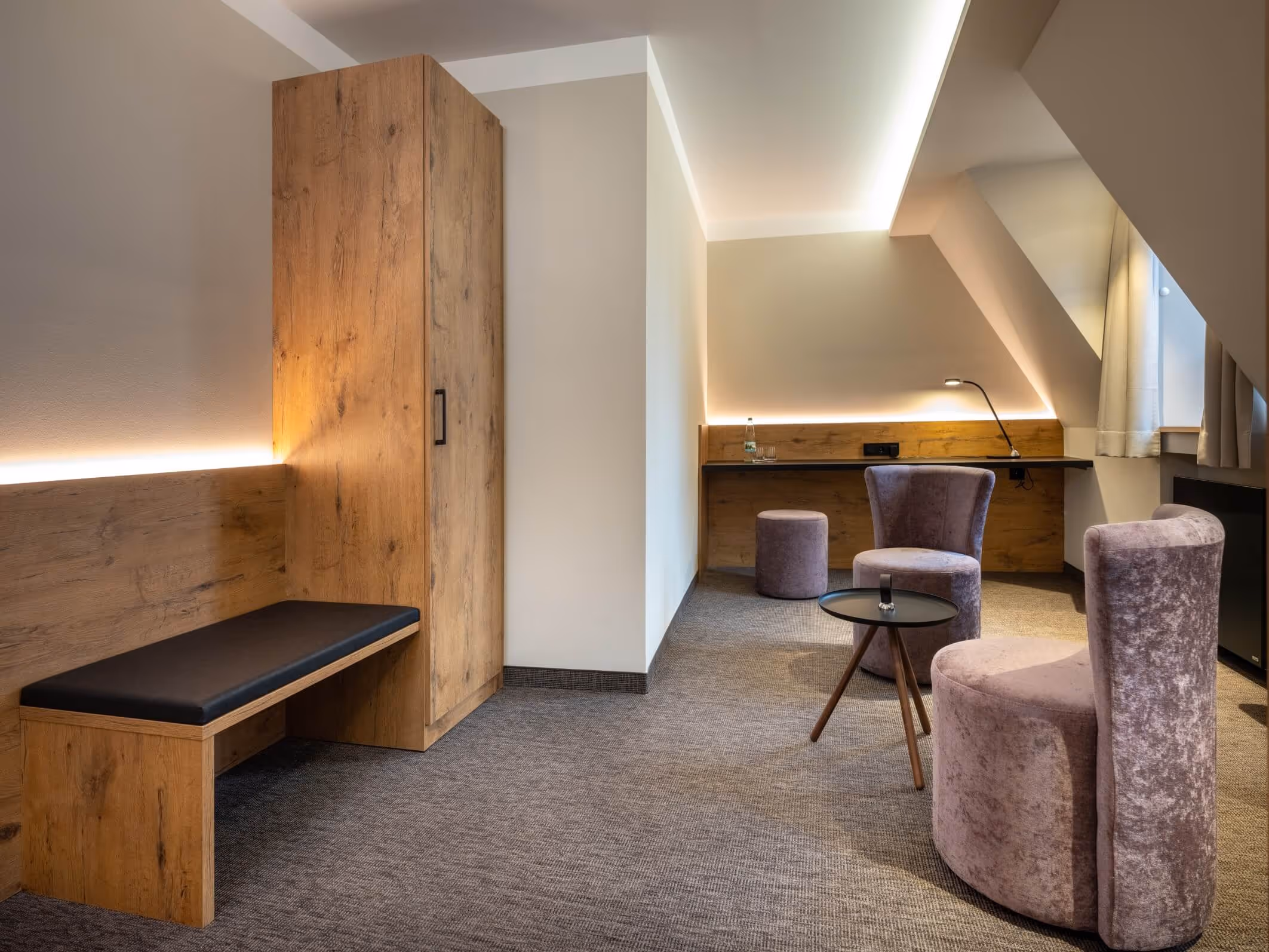 Moderne Hotelzimmerecke im Nashi Rooms Hotel in Erfurt mit lila Samtstühlen, kleinem schwarzen runden Tisch, Holzschrank, Bank und einem Schreibtisch mit Lampe.