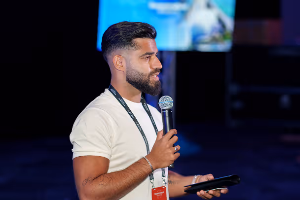 Speaker on stage holding microphone and tablet, wearing white t-shirt and lanyard, with presentation screen in background