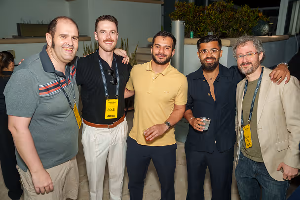 Five male attendees posing together smiling at outdoor evening rooftop event with yellow lanyards