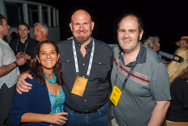 Three attendees posing together at outdoor evening event with lanyards, socializing in background