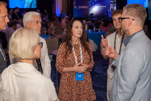 Group of 5 attendees networking in conversation at conference session, blue-lit ballroom with presentation screens in background