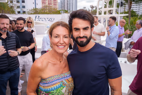 Two attendees posing on yacht deck with city skyline and socializing guests in background