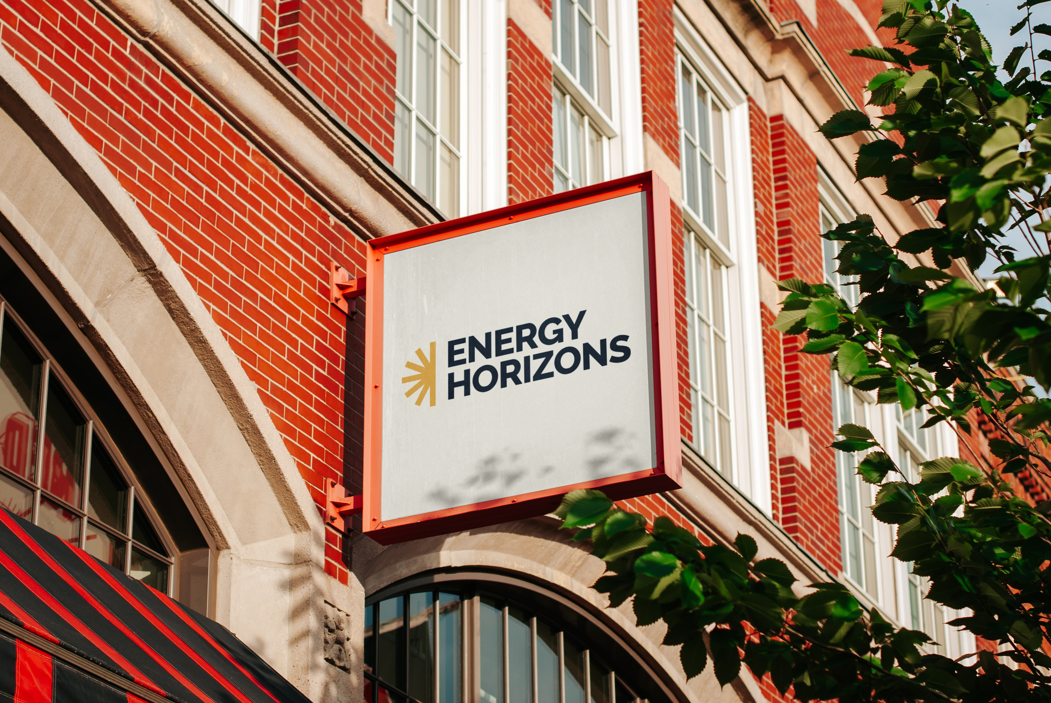 Square sign with red border reading Energy Horizons on a red brick building near green leaves.