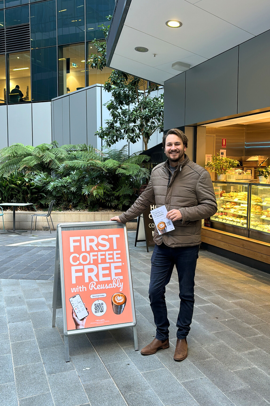 Smiling man in brown jacket holding flyer beside a sign offering a free first coffee with Reusably app.