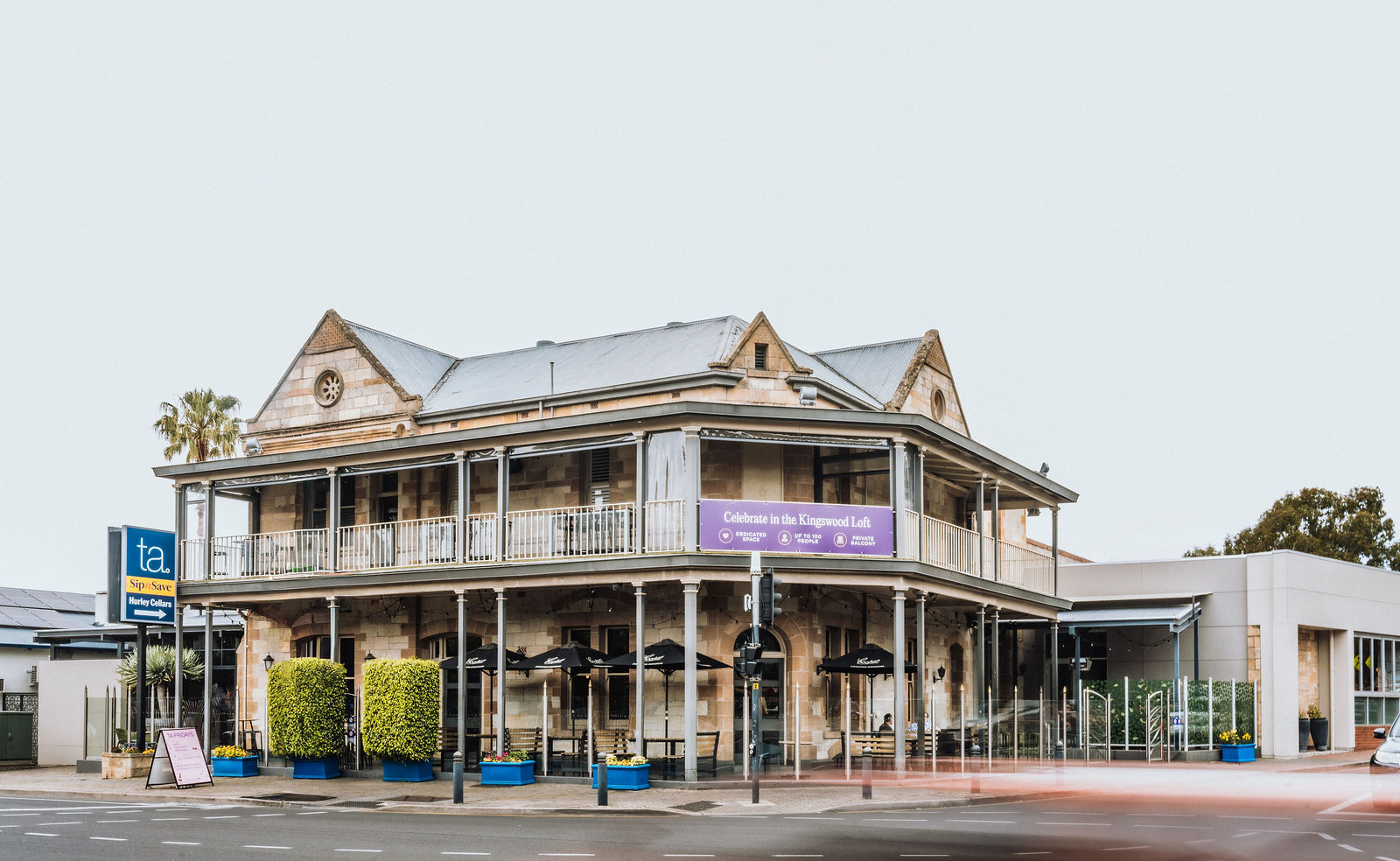 Two-story stone building with a wraparound balcony and outdoor seating at street corner.