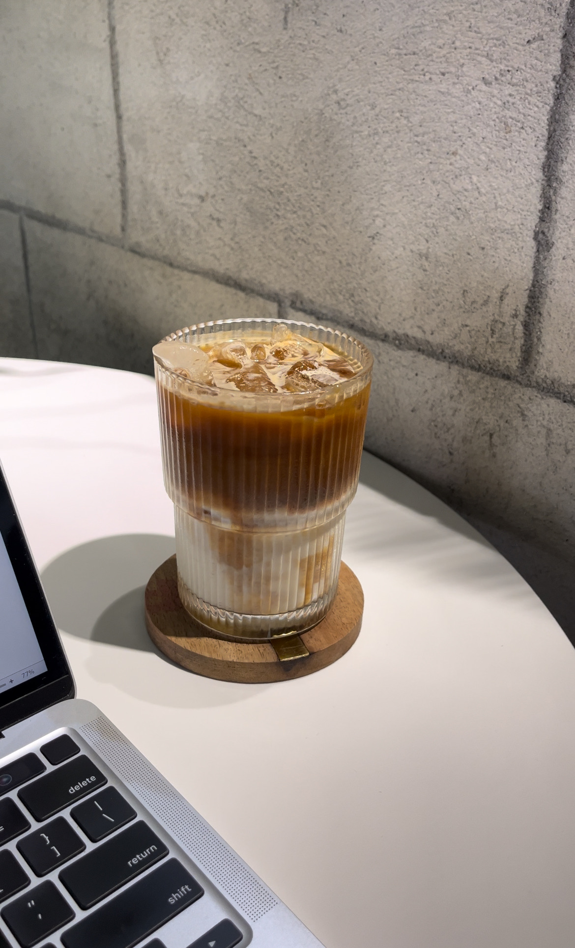 Iced latte with layered milk and coffee in a ribbed glass on a wooden coaster next to a laptop.