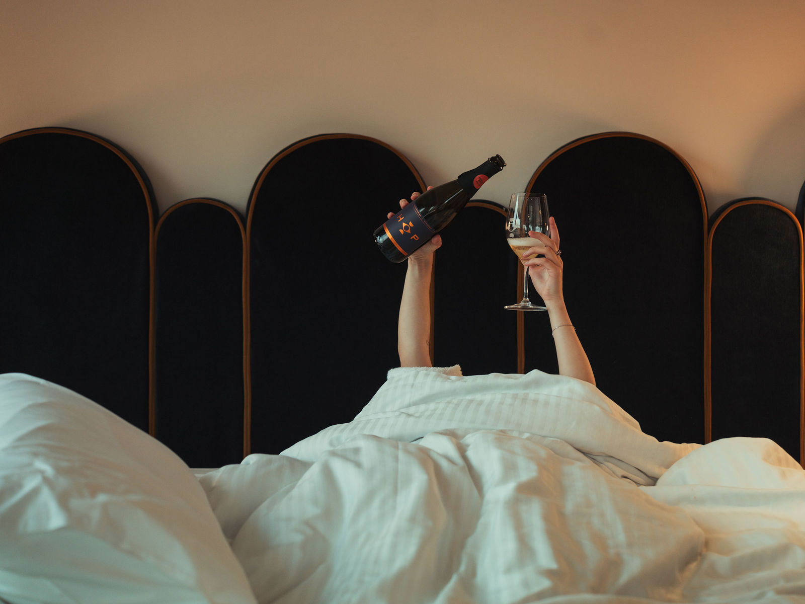 Person under white bed covers holding a champagne bottle and filled wine glass above the bed.