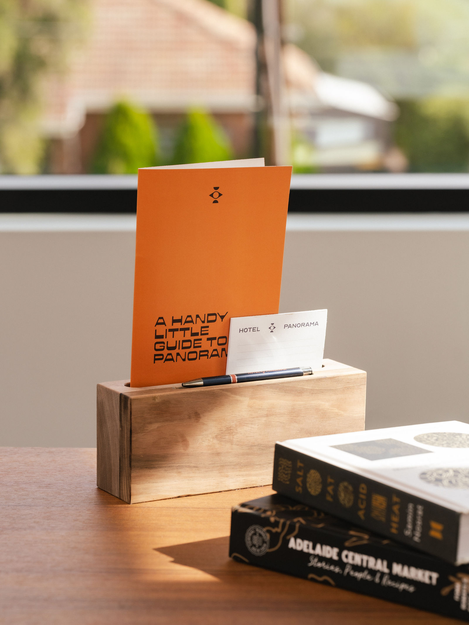Wooden holder on table with orange booklet, hotel card, pen, and two stacked books.