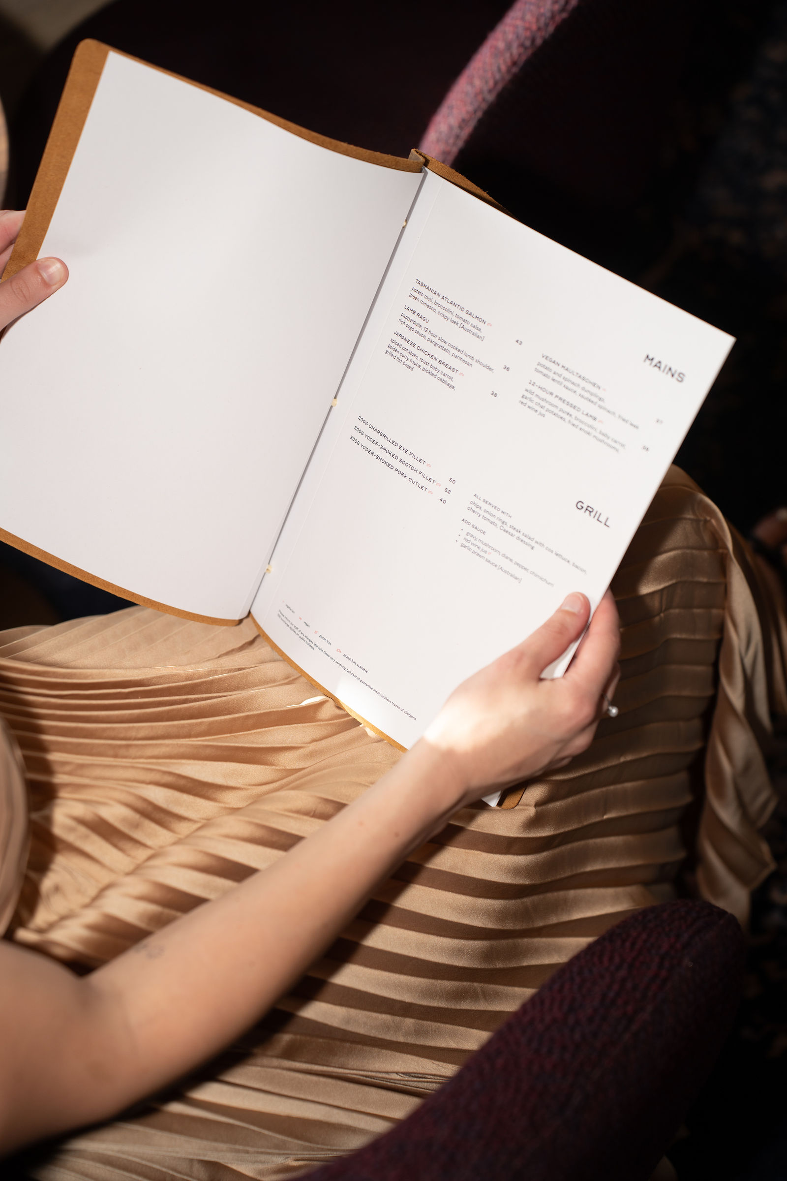 Person holding a menu open to mains and grill sections, sitting on a pleated beige skirt.