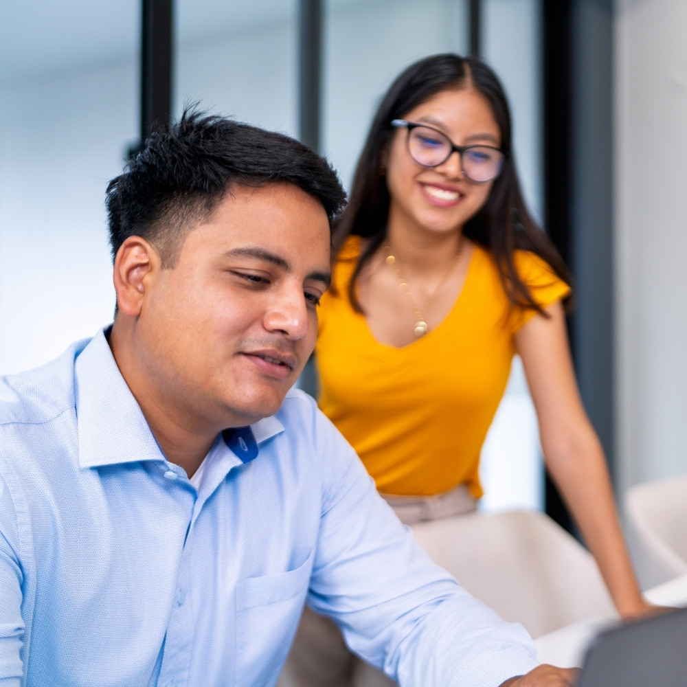 Man in a blue shirt and woman in a yellow top smiling and working together at a desk with a laptop.