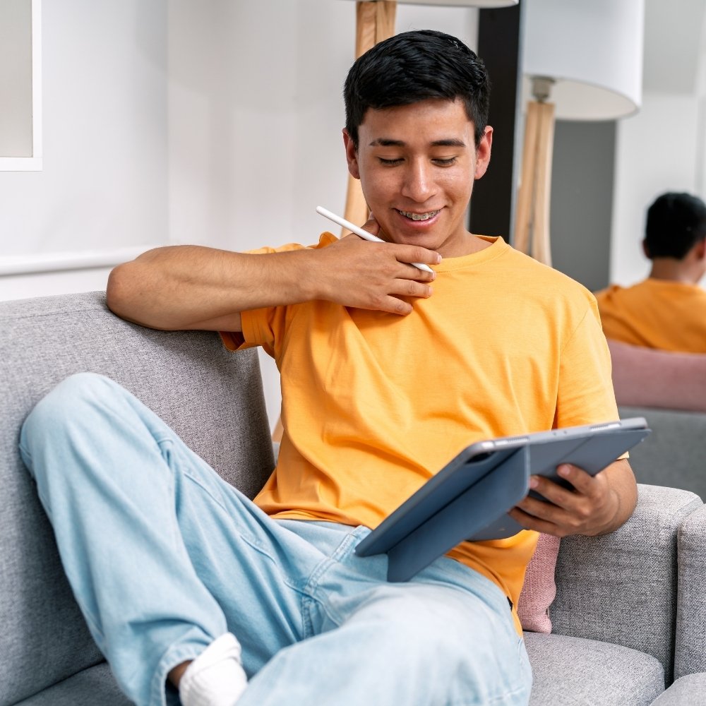 Young man with braces in yellow shirt sitting on couch using a digital tablet and stylus.