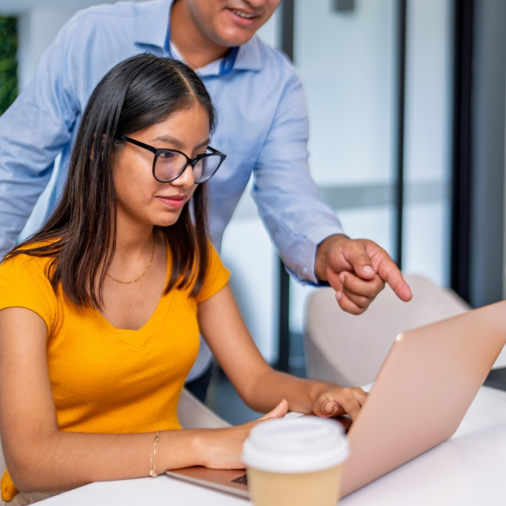 Woman in yellow shirt working on a laptop while a man points at the screen beside her.