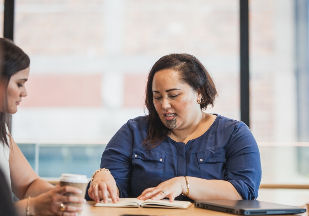 Two women sitting at table, one reading a book and the other holding a coffee cup.