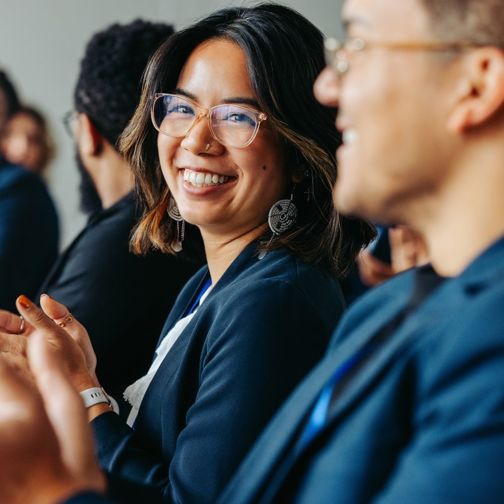 Smiling woman with glasses clapping among several people in a formal setting.