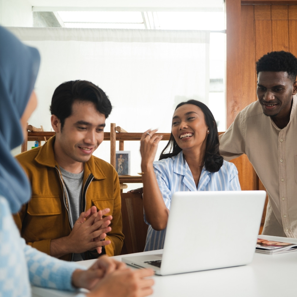 Four diverse people smiling and collaborating around a laptop in a bright office space.