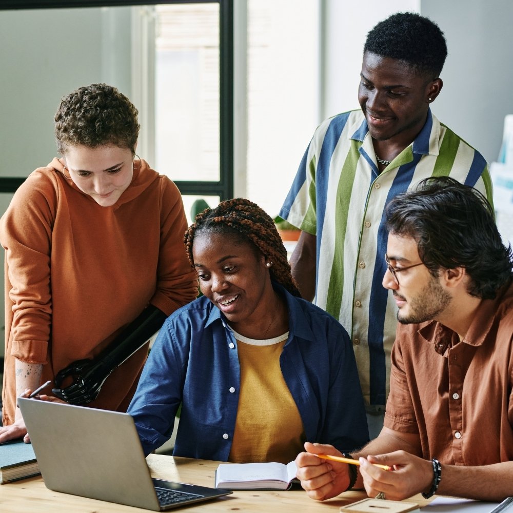 Diverse group of young adults gathered around laptop, collaborating on a project at a wooden table.