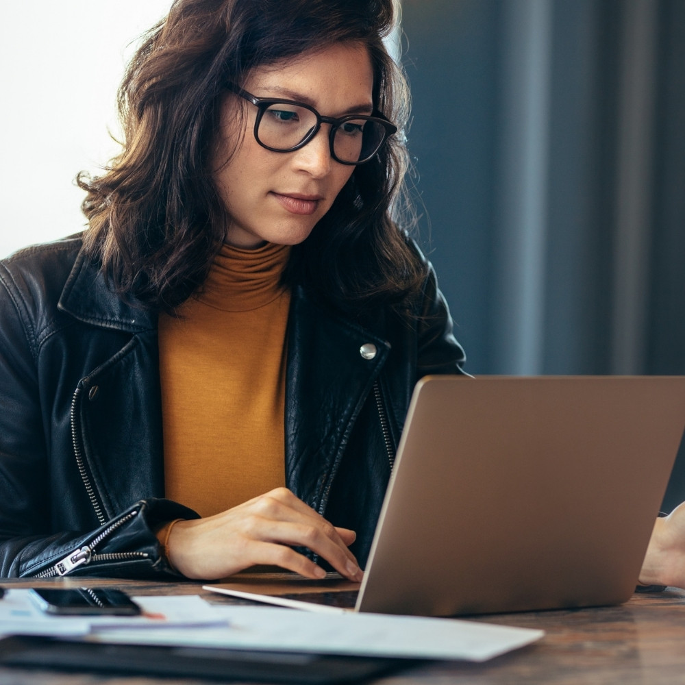 Woman in glasses and leather jacket working on a laptop at a desk with papers and phone.
