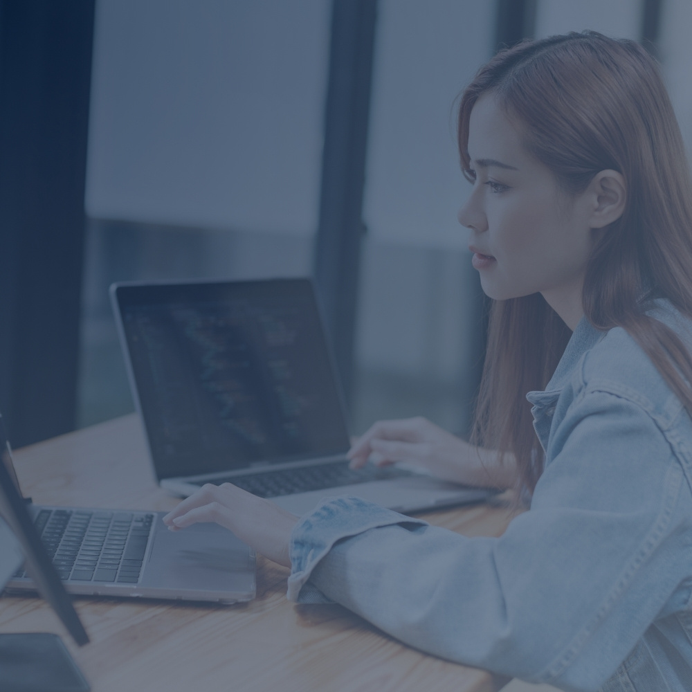 Woman in denim jacket using two laptops side by side at a wooden table.