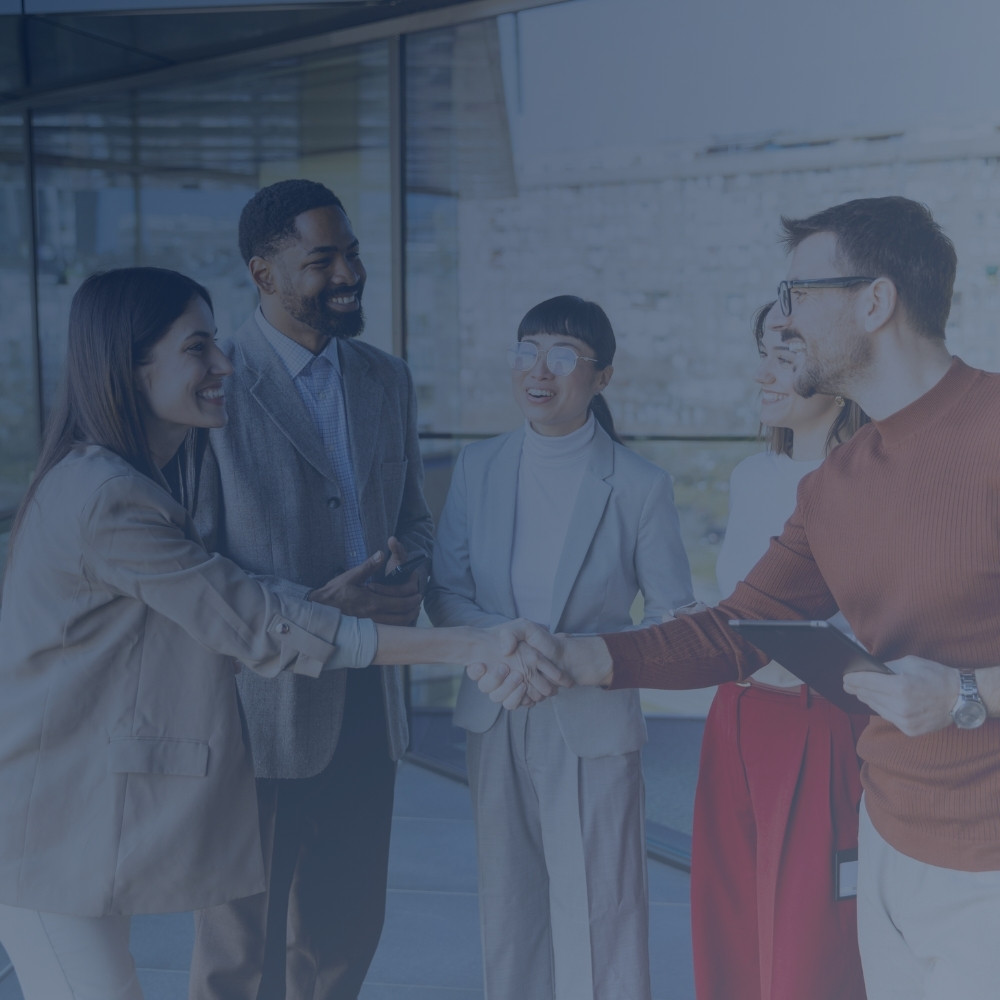 Two professionals shaking hands while three colleagues smile and watch in a modern office lobby.
