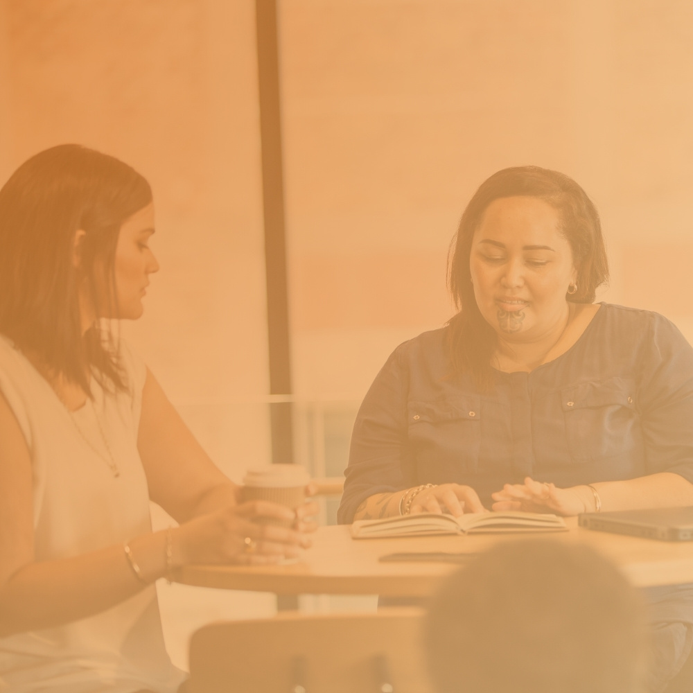 Two women sitting at a table, one reading a book and the other holding a coffee cup.