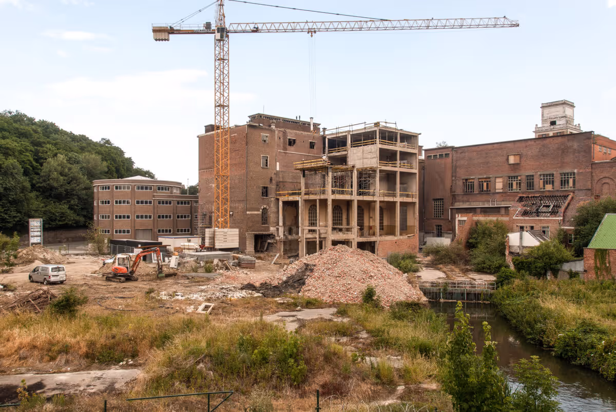Bouwterrein met hijskraan, graafmachine, puinhoop en gedeeltelijk gesloopt fabrieksgebouw langs rivier.