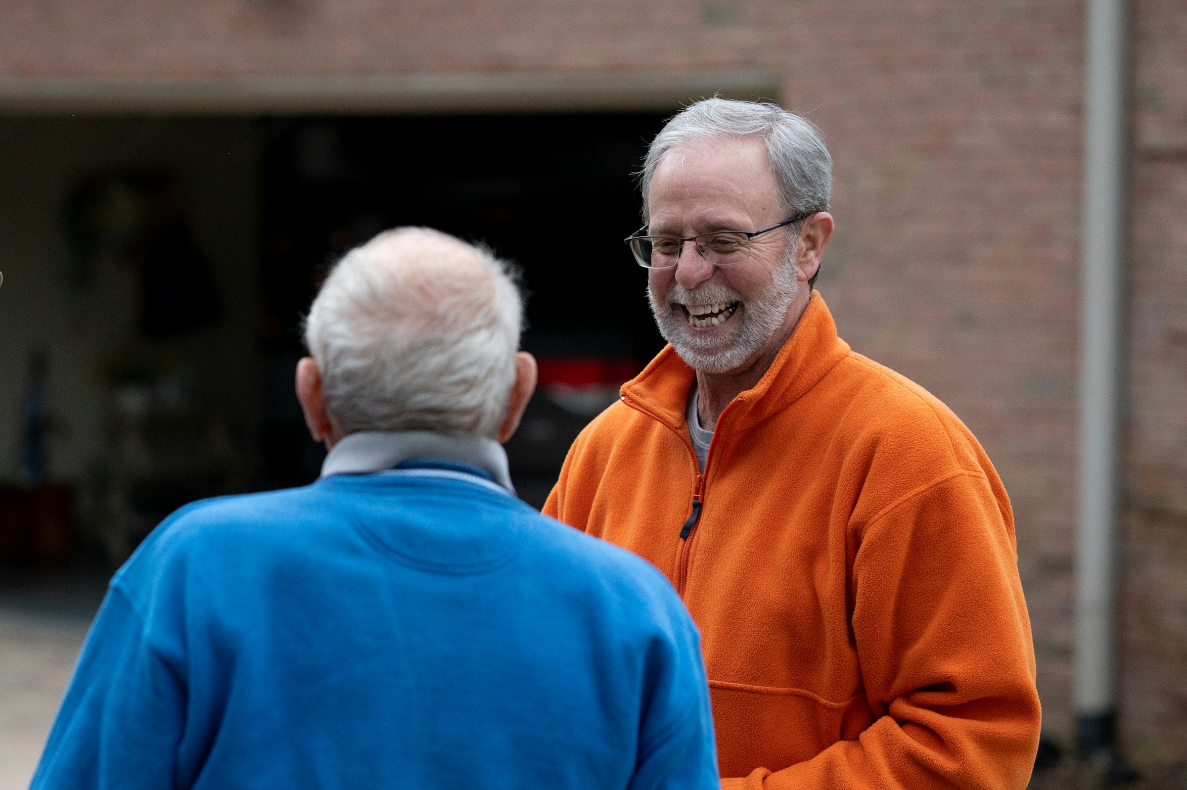 Twee oudere mannen die buiten praten, een lacht terwijl hij een oranje trui draagt.