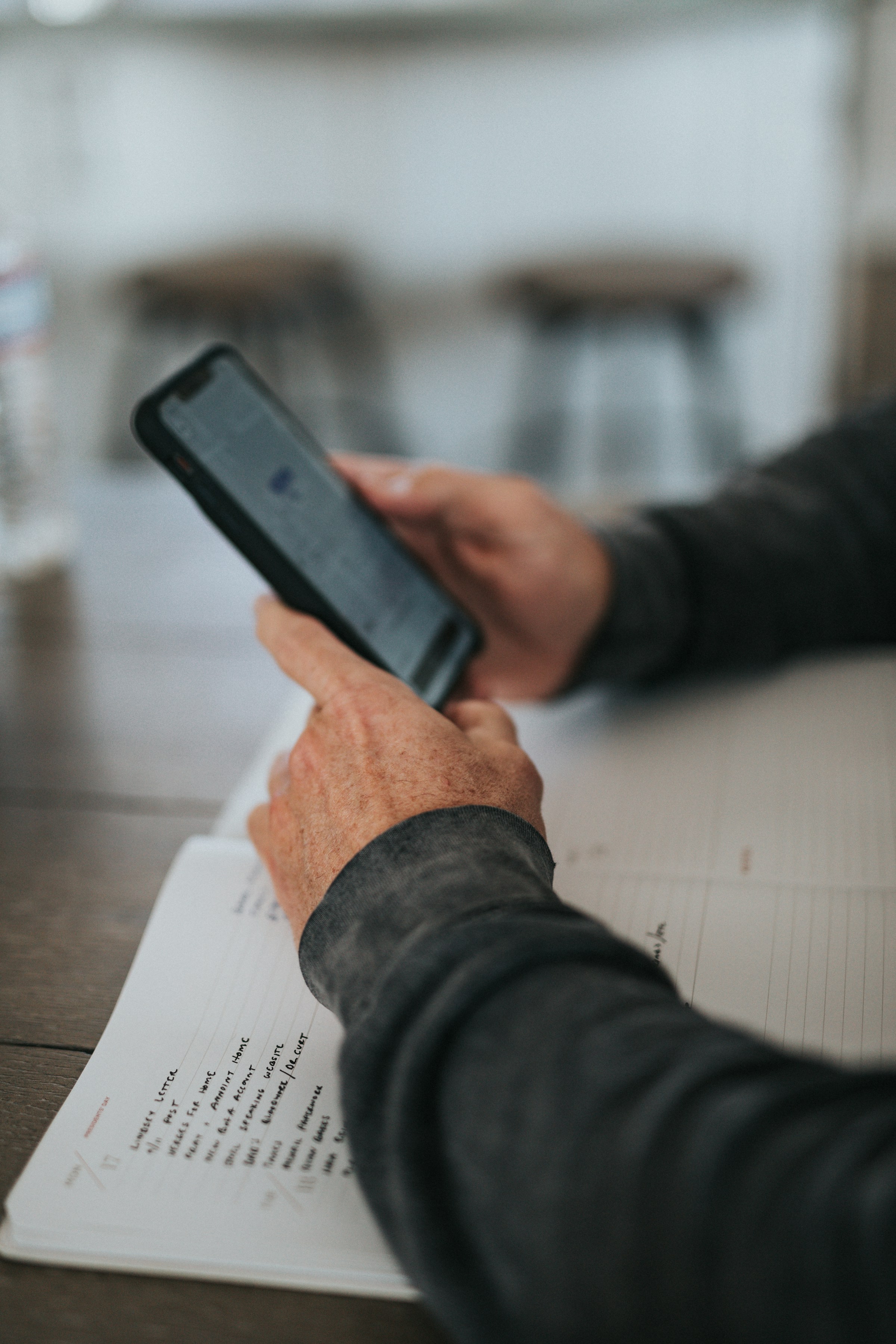 Iemand houdt een smartphone vast en zit aan een tafel met een open notitieboek.