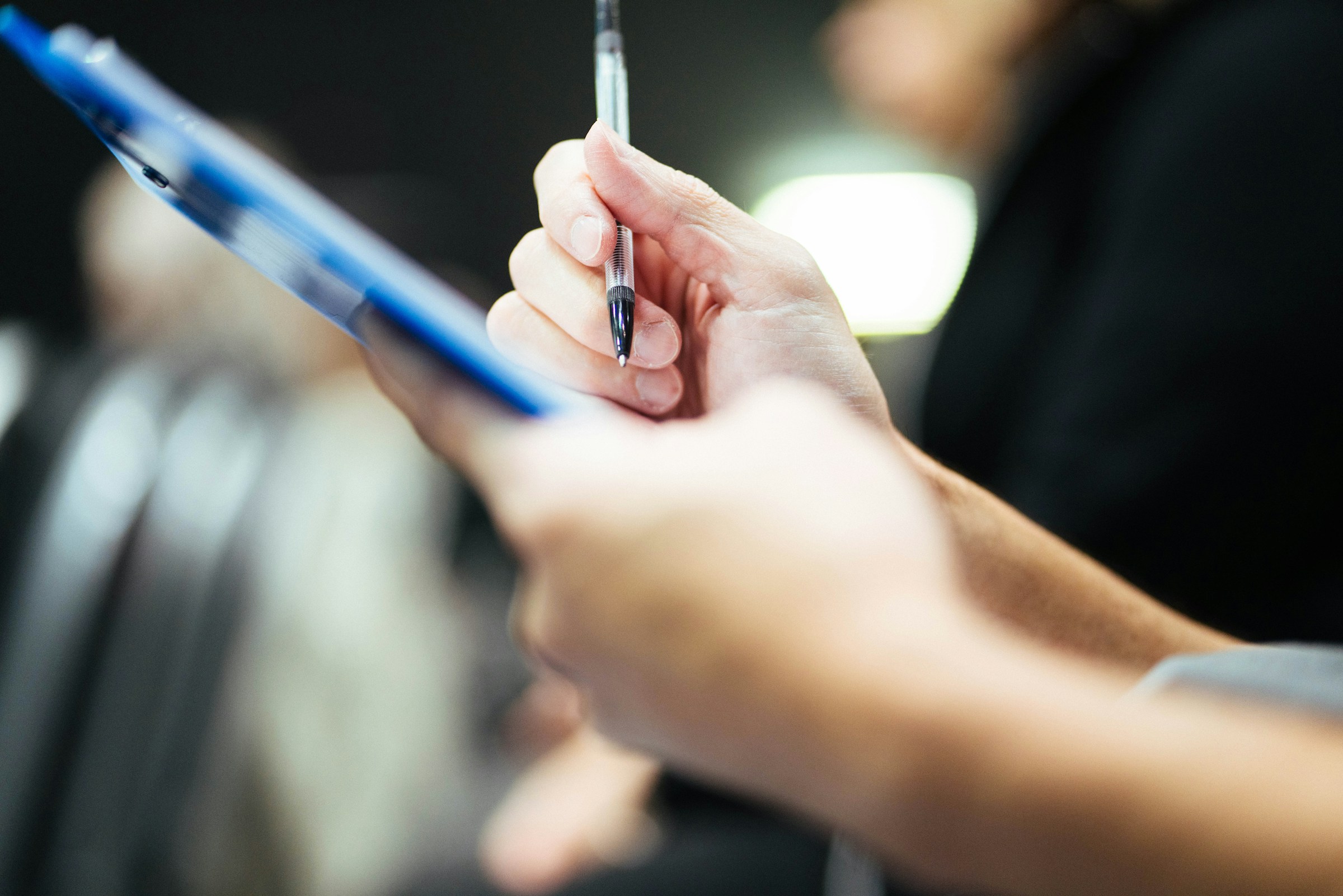 Hand holding a pen and writing on a blue clipboard with blurred background.