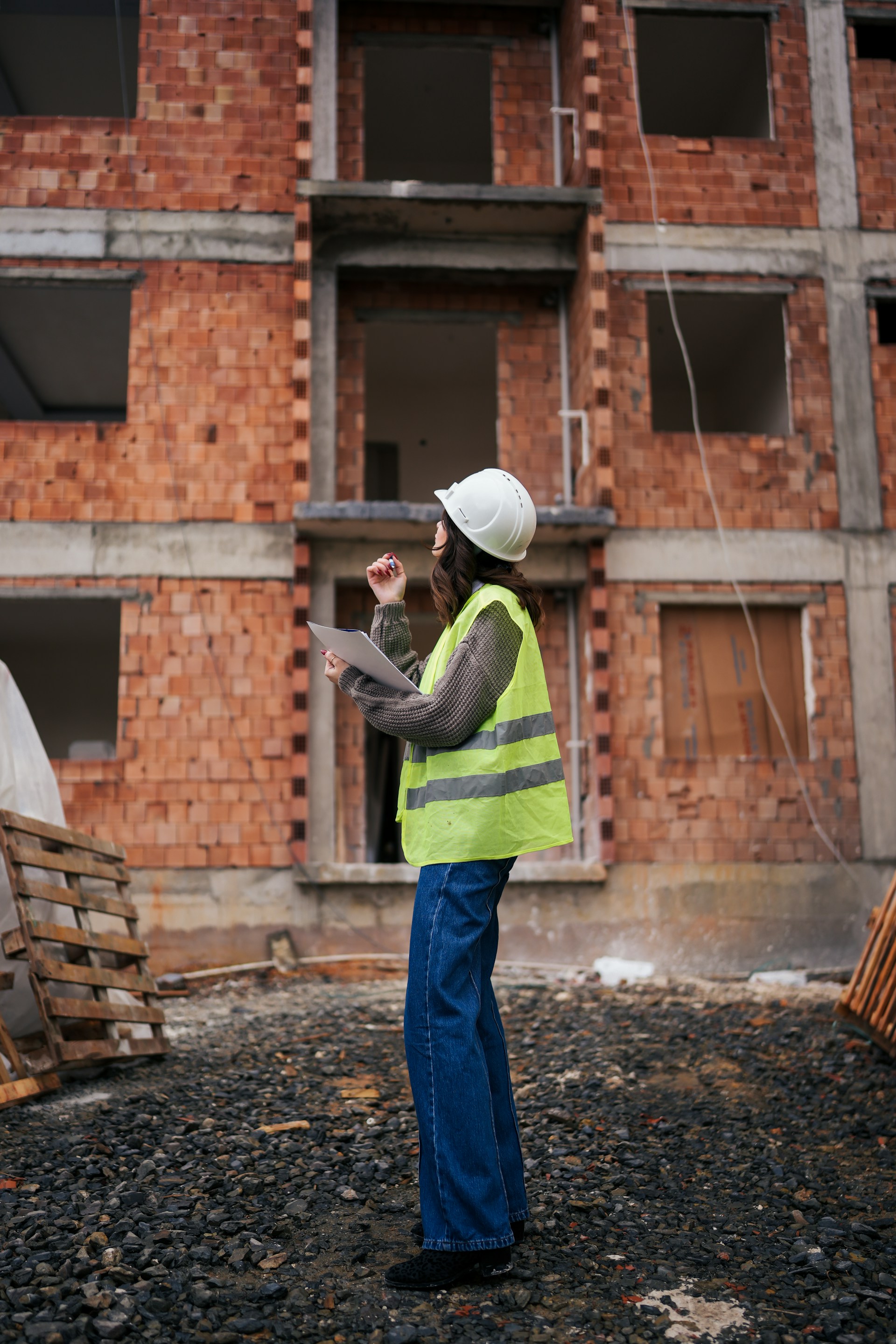Vrouw met bouwhelm en reflecterend vest inspecteert bouwplaats met onafgewerkt gebouw op achtergrond.