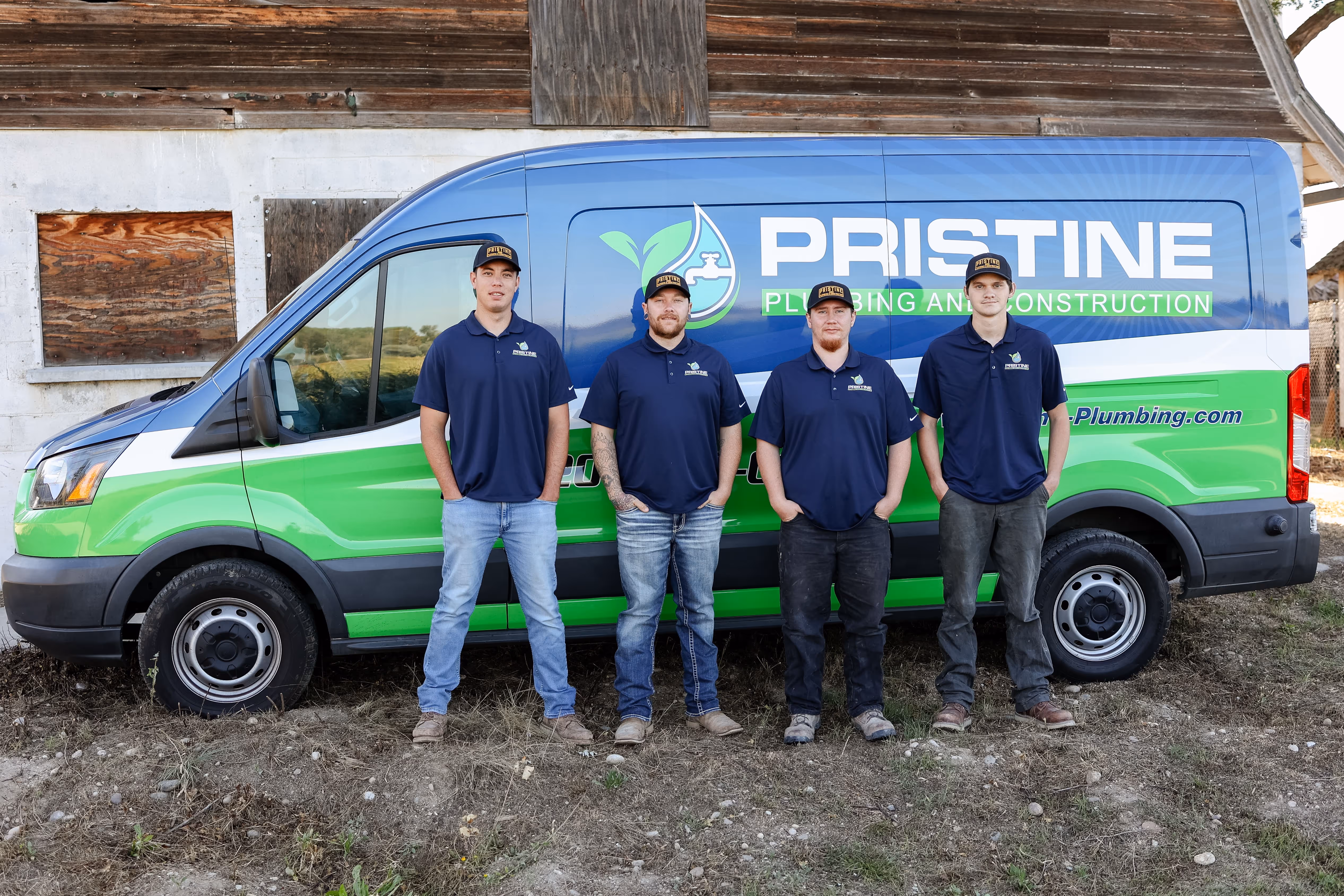 Four men wearing navy blue shirts and caps stand in front of a blue and green Pristine Plumbing and Construction van.