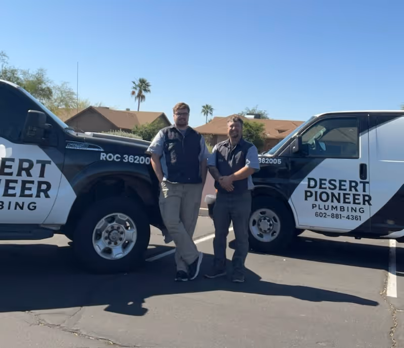 Two men in work uniforms standing in front of two Desert Pioneer Plumbing service vehicles in a sunny parking lot with palm trees and houses in the background.