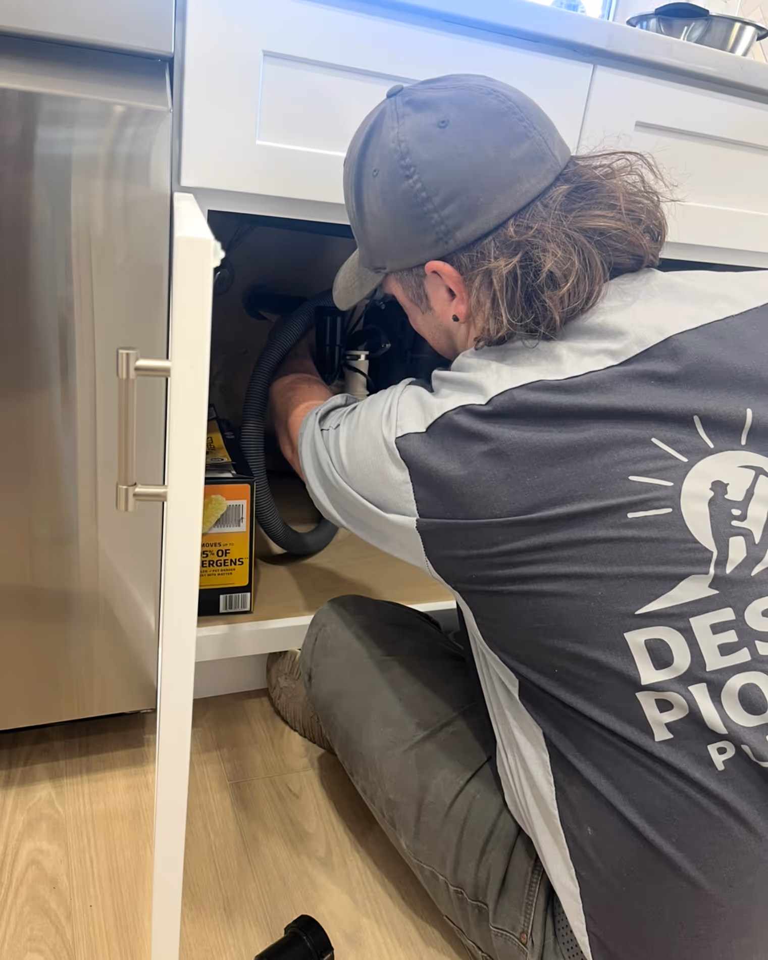 Technician in a cap and uniform working under a kitchen sink cabinet on plumbing or garbage disposal.
