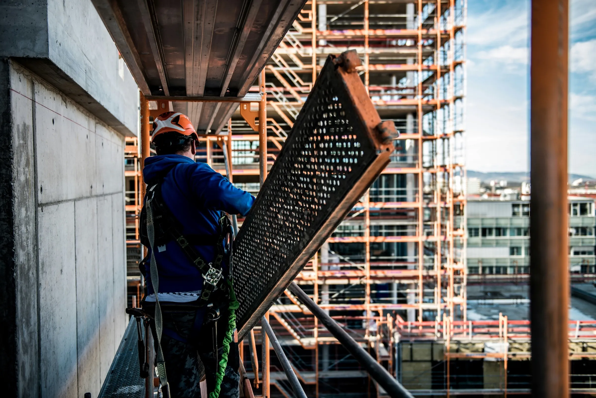 Construction worker wearing safety gear handling a metal scaffolding plank at a building site.