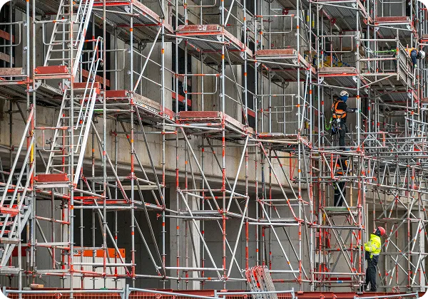 Construction workers on scaffolding at a building site with metal framework and concrete walls.