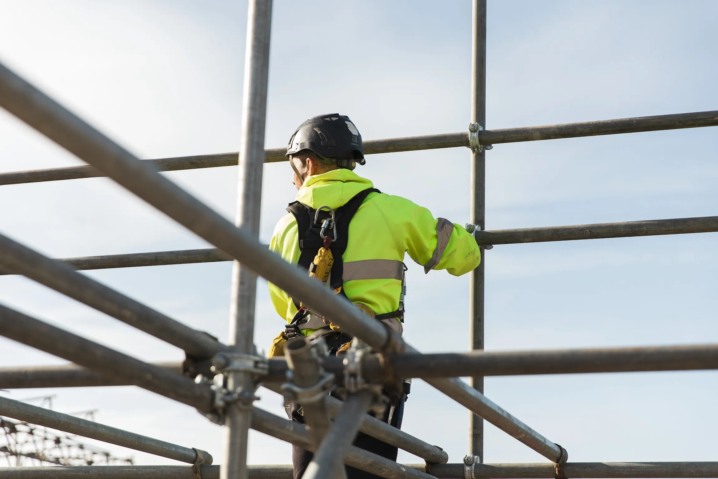 Construction worker wearing a safety harness and helmet assembling metal scaffolding against a clear sky.