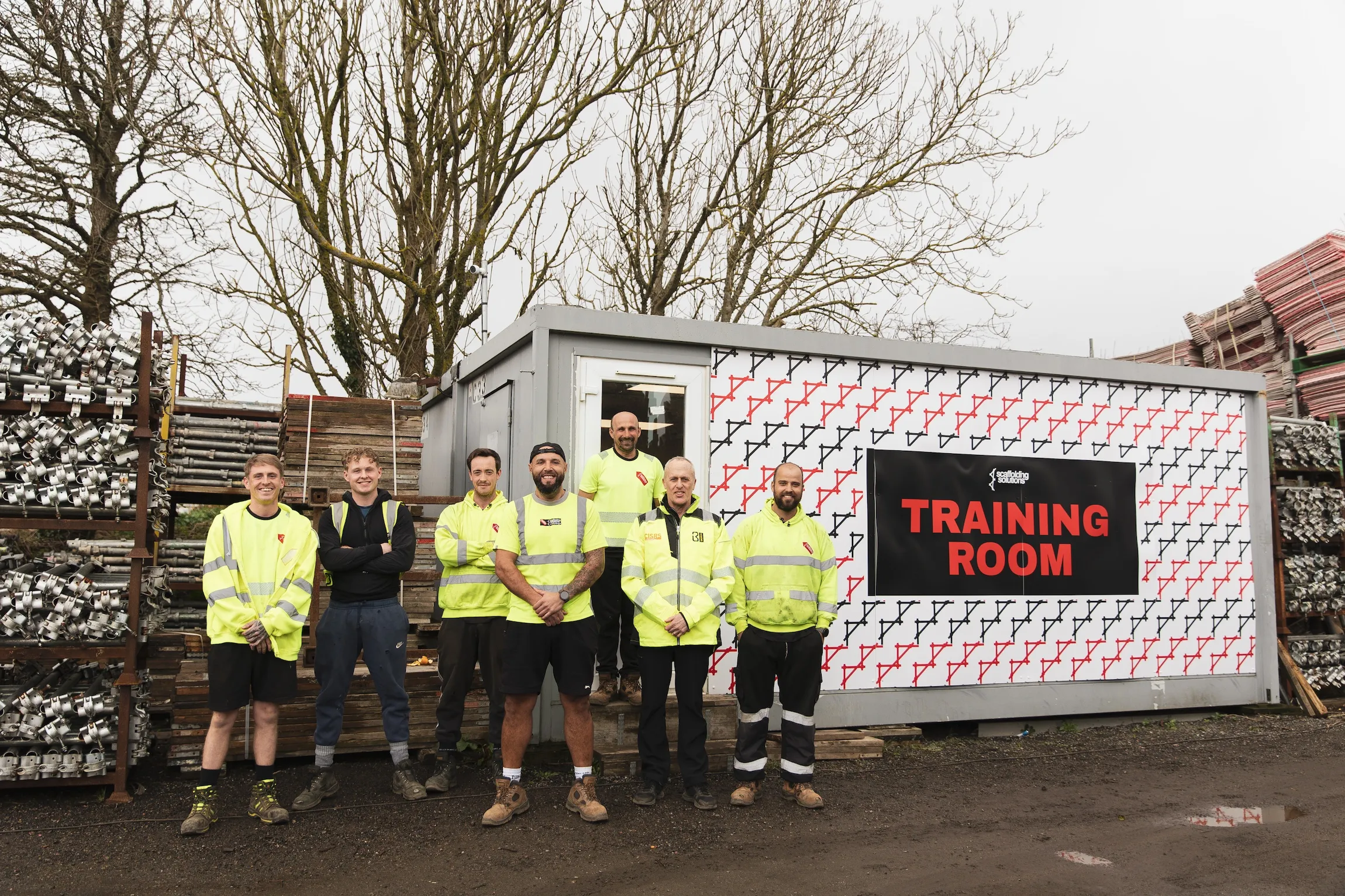 Seven construction workers in high-visibility clothing standing outside a container labeled 'Training Room' at a scaffolding site.