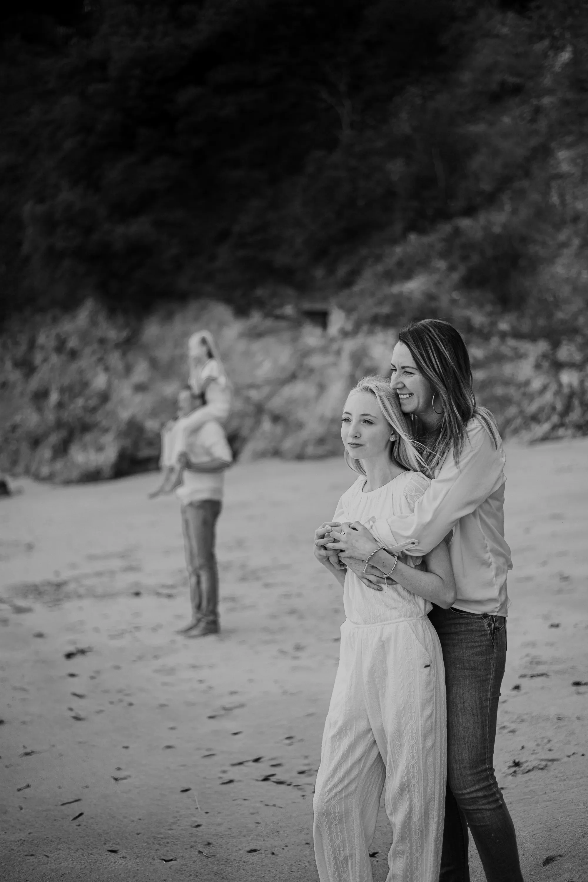 Photographe famille Loire-Atlantique — focus mère et fille se faisant un calin, et en fond le papa qui porte sur ses épaules son autre enfant