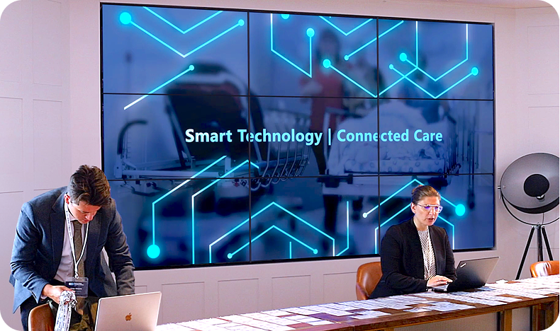 Two people working on laptops in front of a screen showing 'Smart Technology | Connected Care'