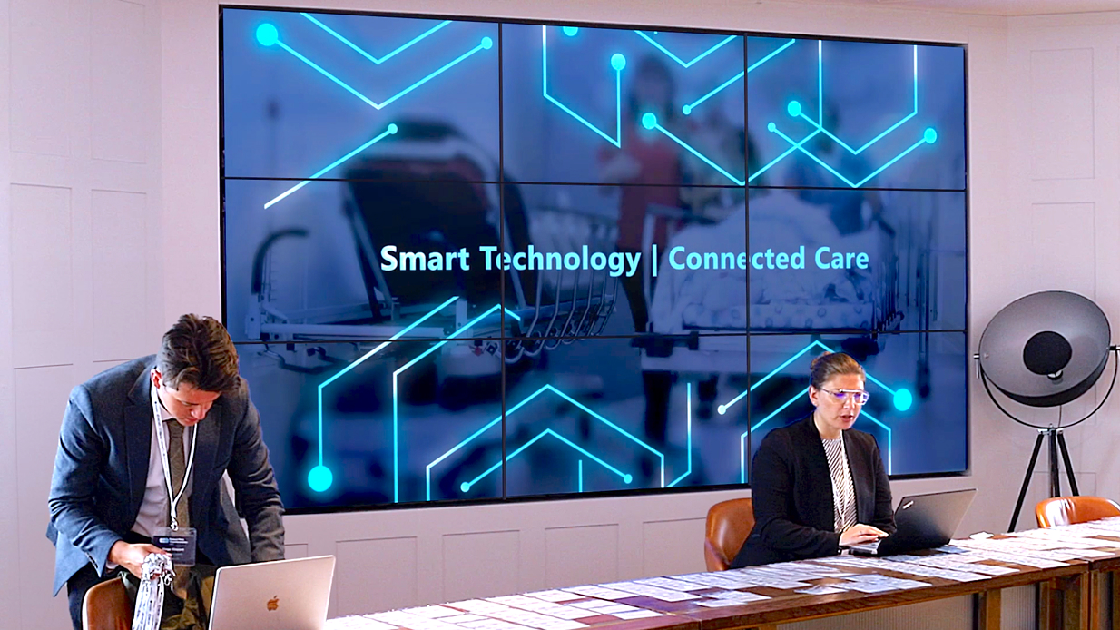 Two professionals working on laptops in front of a screen displaying Smart Technology | Connected Care.