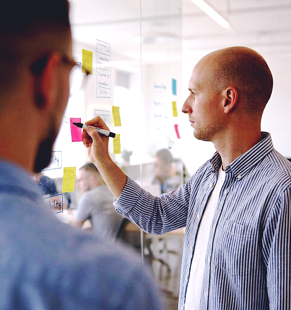 Man in striped shirt writing on sticky notes on a glass wall in a busy office setting.